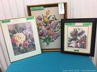 Three framed floral artworks side by side including needlework, oil painting by Herman Pils, and watercolor by Lorraine Heryet.