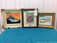 Three framed paintings displayed side by side on a blue surface against a white wall.