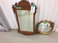 Photo showing both mirrors side by side. One large rectangular wood-framed mirror standing upright with decorative top and bottom cutouts. Second smaller oval mirror with a gold ornate frame placed on the floor.