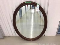Front view of the oval mirror leaning against a storage unit door, showing the full frame and reflective glass.