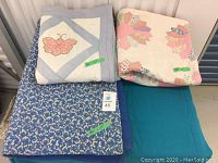 Three handmade quilts folded and stacked. Quilt on top has butterfly appliqué and lattice design. Middle quilt shows cream fabric with multi-colored flower shapes. Bottom quilt is blue with small white flowers.