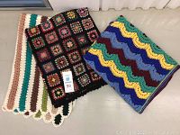 Three handmade crocheted afghans arranged on floor. One folded with visible striped scalloped edge design, one with granny square colorful design on black, one folded with wavy stripes pattern.