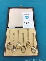 Top view of the open fitted box housing six small sterling silver spoons laid flat on a cushioned beige base, with a satin-like cushioned beige lid interior. A card indicating lot number and spoon size at 4 inches is present.