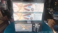 Three boxed ceiling fans stacked on a table: two 42-inch SMC decorative ceiling fans and one 52-inch Peachtree Choice ceiling fan.