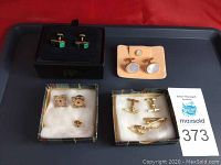 Four pairs of men's cuff links displayed on a tray, each pair in or near small presentation boxes with padded inserts. The cuff links vary in style and material.