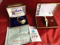 Both watches displayed side by side with boxes and paperwork on red cloth backdrop