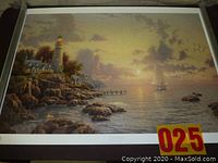 Full view of the framed lithograph depicting a seaside scene with lighthouse, sailboat and rocky shore at sunset.