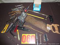 Lot of various hand tools arranged on a table. Front center shows a rusty handsaw with wooden handle, small hacksaw, assorted wrenches, utility knives, screwdriver, and a box of screws.