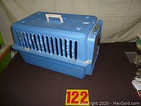Blue plastic pet carrier with handle, ventilation slots, and metal mesh door, placed on a dark surface with seller tag number 122.
