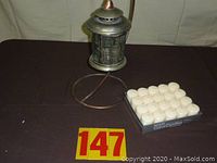 Full view showing the metal lantern on the circular stand and the package of 20 white votive candles nearby.