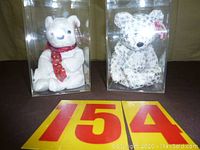 Two white Beanie Baby bears displayed in clear plastic cases; one plain with red star scarf, one with shiny star designs all over