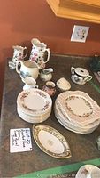 Photo showing assorted bone china plates, creamers, and urns arranged on a kitchen counter, including stacks of dinner and bread plates, various creamers, a candy dish and urns