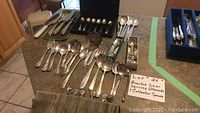 Full spread of silver utensils on countertop including serving spoons, collector spoons, carving set in velvet roll and presentation cases