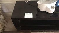 Full view of the black TV stand showing the top surface and glass shelves below. Includes a label showing the lot number and measurements.