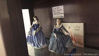 Front view of two Royal Doulton figurines on display shelf, showing details of dresses and poses.