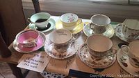 12 bone china tea cups with matching saucers displayed on table showing assorted floral designs and colors including pink, green, white, and gold trim.
