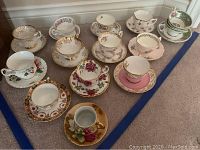 Overview of all 13 bone china tea cups and matching saucers arranged on carpet surface, showing various floral and gold trimmed designs.