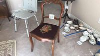 Photo of first wooden chair with carved top rail and colorful floral needlepoint seat on dark brown background, accompanied by teacups and other household items nearby.
