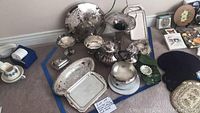 Overview of the assorted silver plated serving items on a carpet floor next to some household objects.