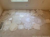 Photo showing an assortment of vintage white doilies in different sizes and shapes arranged on a carpeted floor near a window and vent.