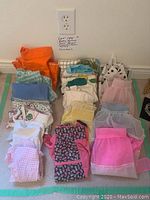 Top-down photo showing the collection of 16 colorful vintage women's aprons laid out with some folded, featuring bright colors including orange, pink, blue, floral patterns, and sheer fabrics with lace trim.