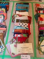 Photo shows multiple stacks of folded silk scarves laid out on a carpet floor with green tape lines creating sections. The scarves vary in color and pattern, including florals, polka dots, solid borders, and abstract prints.