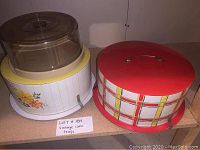 Three cake trays including two metal vintage trays with colorful designs and clips and one glass tray with clear dome lid.