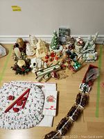 Photo showing Christmas placemats, candles, napkins, bell ornaments, decorative buildings, sleighs with horses, and caroller figurines arranged on the floor.