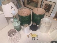 Photo of two vintage lamps, assorted lamp shades of different sizes and colors, displayed on carpeted floor against a wall with framed pictures.