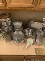 Wide view showing a variety of metal baking pans, bowls, a sifter, and utensils on a kitchen counter below wooden cabinetry