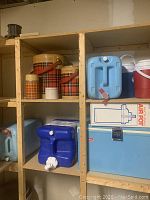 Shelf holding vintage plaid thermoses, blue water jugs, red and white water cooler jugs, and a Coleman cooler box