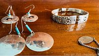 Photo shows two pairs of sterling silver earrings, one with turquoise beads and round discs, the other with horse silhouette cutouts, alongside a sterling silver bracelet with detailed floral design.