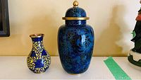 Two cloisonne decor pieces shown together on a shelf: smaller blue and yellow vase and larger blue lidded jar.