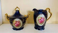 Photo showing teapot with lid and creamer side by side, highlighting deep blue color, gold detailing, and floral medallions.