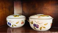 Pair of round covered porcelain casseroles with fruit and vegetable designs, white base color with gold-tone handles on lids and bodies