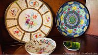 View of four fine bone china items: one round plate with floral/gold gilt design, one deep blue floral plate, one pierced floral bowl, and one small decorative tray.