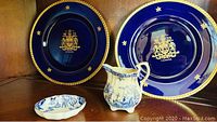 Two large Royal Crown Derby plates in cobalt blue with gold trim, maple leaf designs and a gold Canadian crest, shown together with a Simpsons Pottery cream and blue pitcher and matching dish.
