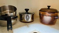 Metal wok with handles, stainless steel pot with lid, stainless steel bowl, and amber glass Visions pot visible on shelf.