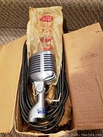 Microphone seated in original cardboard box with coiled cable and branded paper sleeve visible