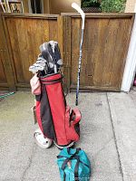 Full view of red and black golf bag with Bob Joski International golf clubs and Reebok hockey stick in the background, Torino bocce set bag on ground.