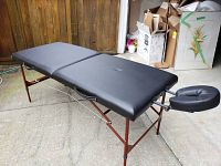 Full view of the black portable massage table set up with headrest attached, standing on wooden legs with cross braces.