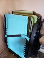 Two outdoor lounge chairs stacked and folded, one light blue and one light green with black metal frames.