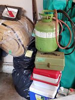 Photo of rolled beige sleeping bags, green propane tank with hose, fishing tackle box, stacked cooler boxes, and two teal camping chairs