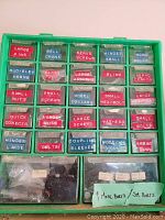 Green plastic compartment organizer with labeled sections containing various small hardware parts including nuts, bolts, screws, washers, hinges, and collars.