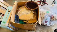 Box containing assorted fabric pieces, wicker basket, and some pillows in white bags.