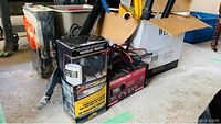 Overall view of boxed headlight restoration kit, Craftsman battery charger, cables, and some metal bars visible on concrete floor.