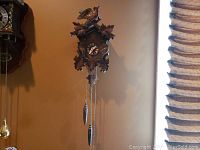 Photo showing one carved wooden cuckoo clock hanging on wall with bird, leaf decorations, Roman numeral clock face, chains and weights.