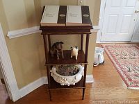 Front view of a dark brown wood podium with three levels showing books on top and decorative items on the lower shelves, placed in a room corner with hardwood floor and beige walls.
