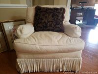 Front view of cream textured upholstered armchair with fringe detailing on bottom and dark decorative throw pillow