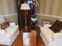 Angle showing gold cube chest positioned between two chairs with white slipcovers, the brass floor lamp behind and to one side.